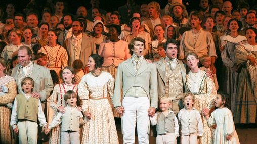 A large group of performers dressed in pioneer clothing on stage singing. 