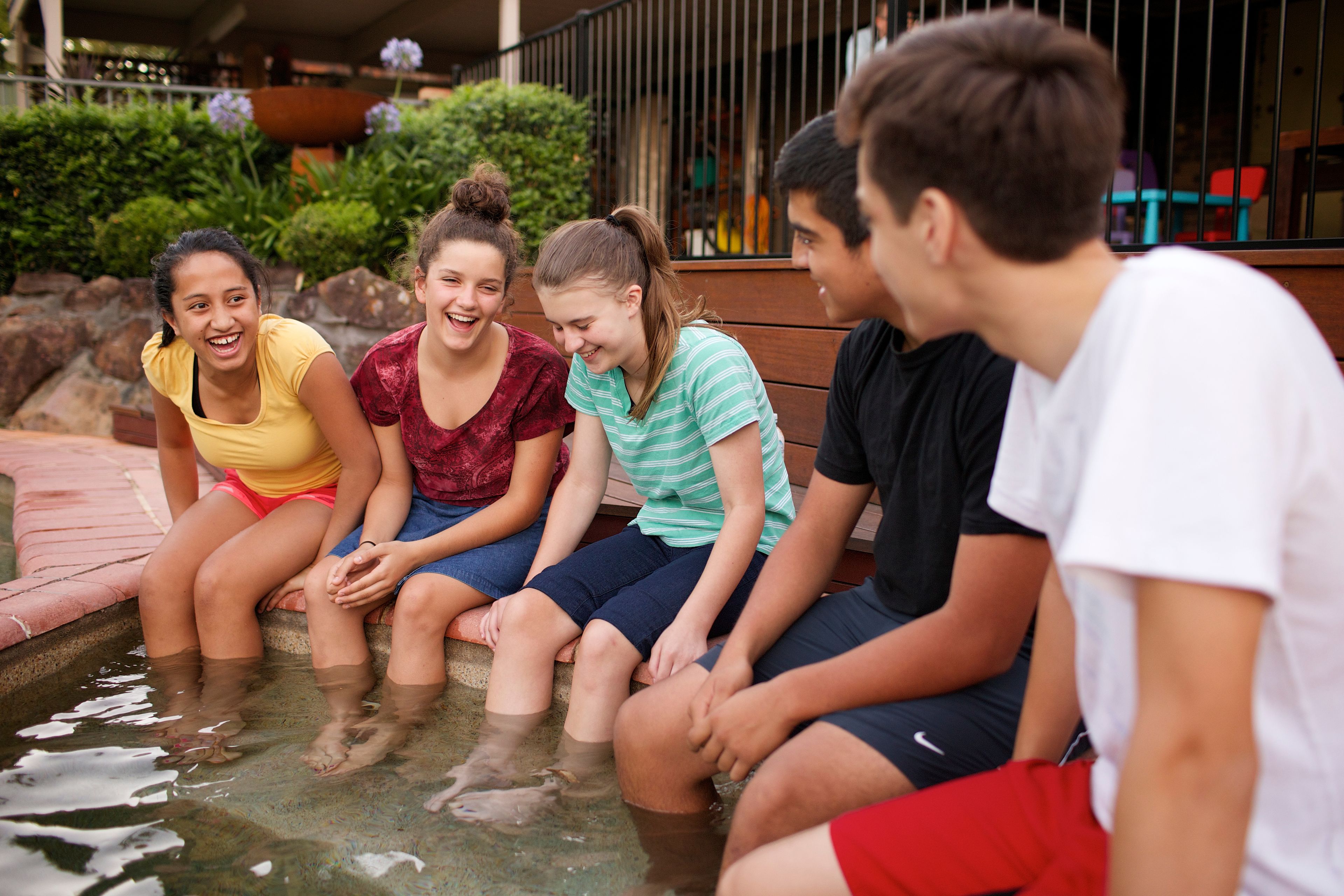 A group of youth hanging out together on the edge of a pool, with only their feet dangling in the water.  