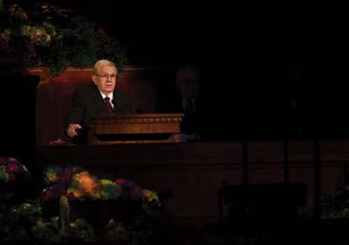 Boyd K. Packer sitting in a red armchair while speaking at the pulpit in a session of general conference.