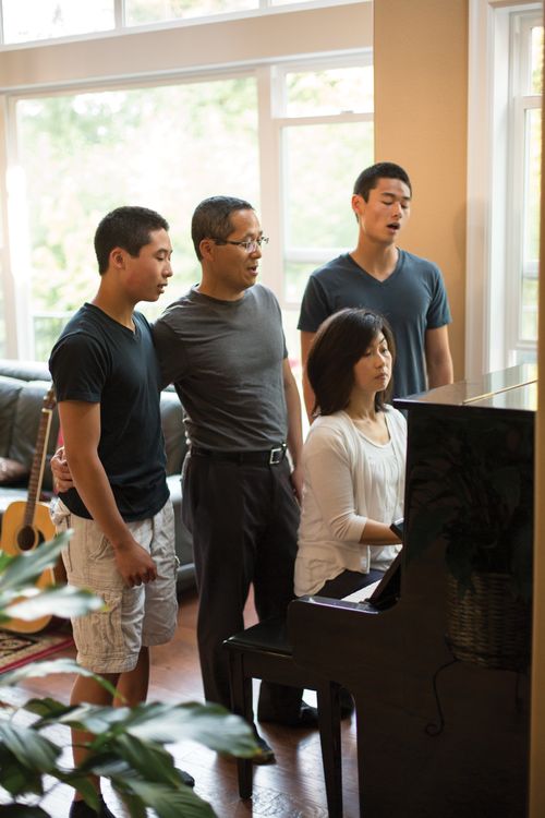 A mother plays the piano in the living room with her husband and two teenage sons standing behind her, singing along.