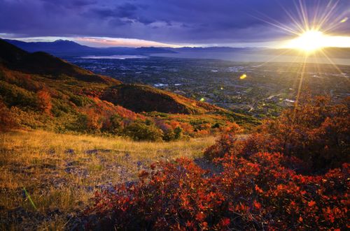 The sun sets behind a valley, with the hills in the foreground covered in changing leaves during autumn.