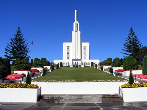 The front of the Hamilton New Zealand Temple, with a green lawn leading toward the doors and interspersed with blooming flower beds.