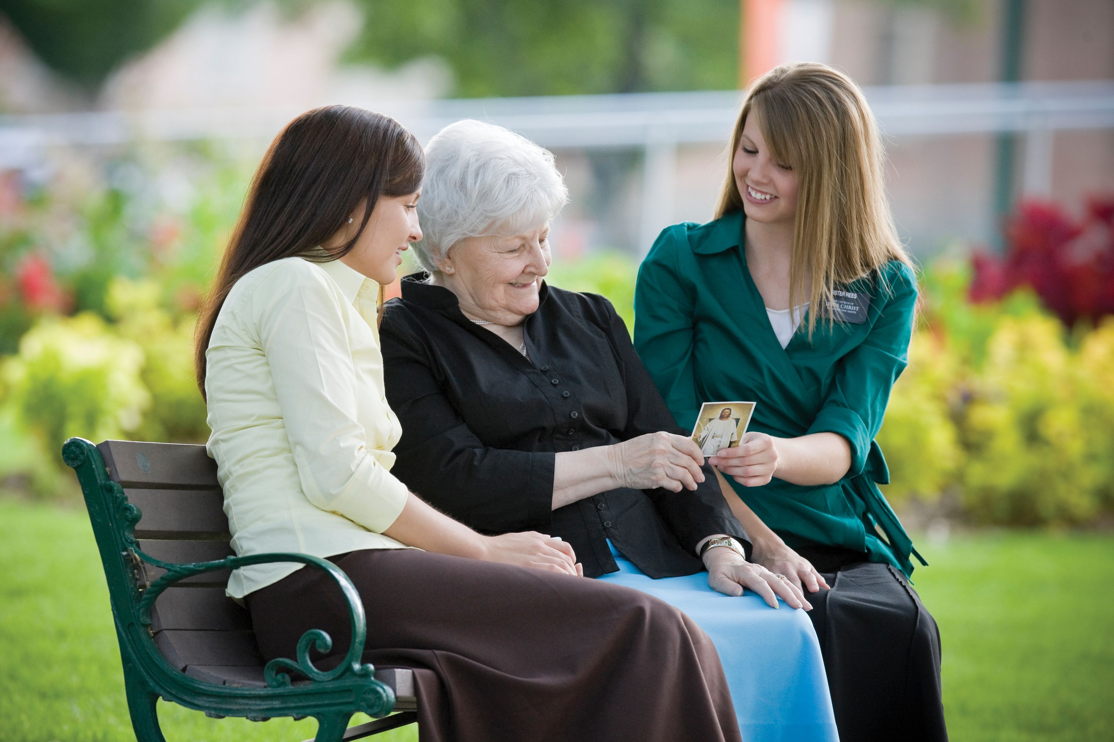 An elderly woman sitting between two sister missionaries and looking at a pass-along card.