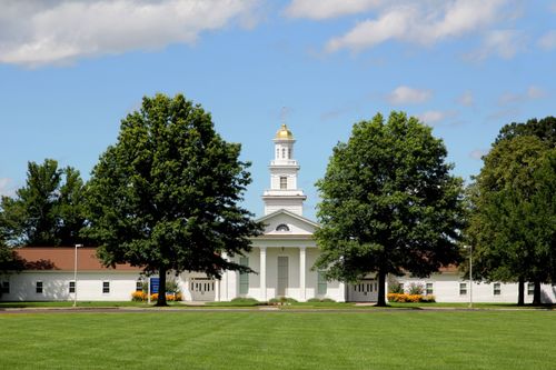 A large grassy lawn and trees in front of a white chapel and visitors’ center with a steeple at Peter Whitmer’s farm in Fayette, New York.