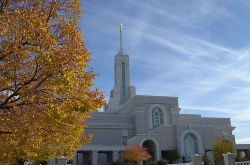 The Mount Timpanogos Utah Temple in the fall, with an orange colored tree seen to the left of the entrance and a blue sky with thin white clouds above.