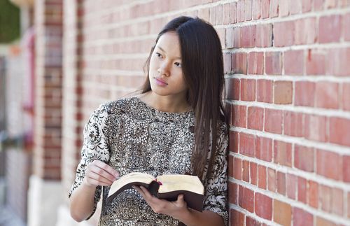 young adult woman holding the scriptures and leaning against a wall