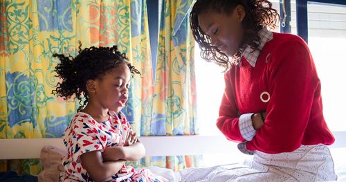 A young woman is kneeling with a little girl on her bed. They fold their arms and pray together. The window is seen behind them.