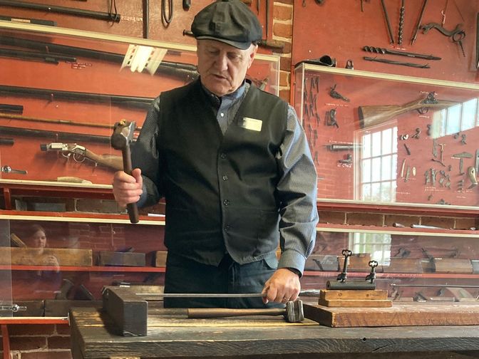 Senior missionary with hammer in hand shapes metal on an anvil. Tools and rifle parts are on display in the background.