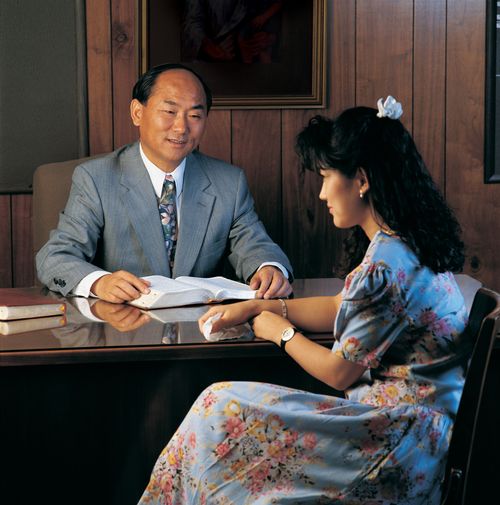 A young woman in a floral print dress sits in front of her bishop, who has a set of scriptures open in front of him.
