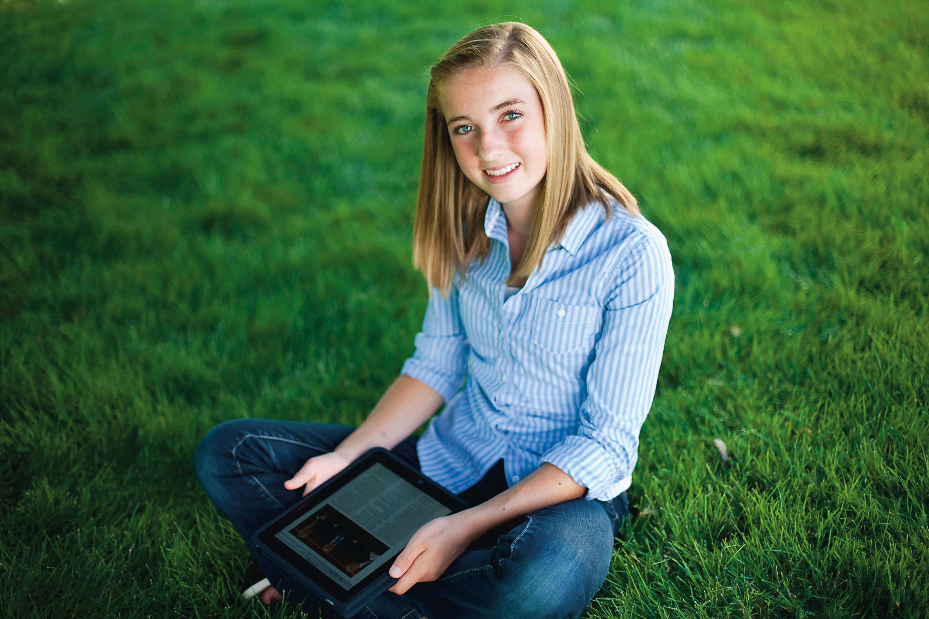 A young woman sits outside on the grass and holds a tablet.