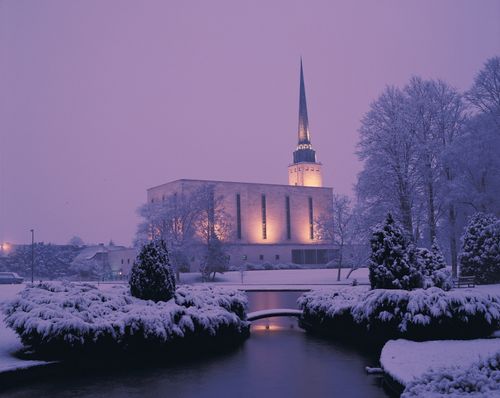 A side view of the London England Temple covered in snow on a winter evening, with the temple pond in the foreground.