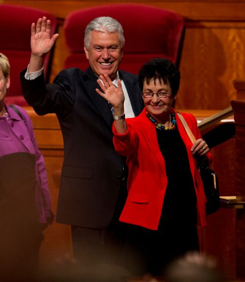 Dieter F. Uchtdorf in a black suit and Harriet Uchtdorf in a red blazer, standing and waving to the congregation in the Conference Center.