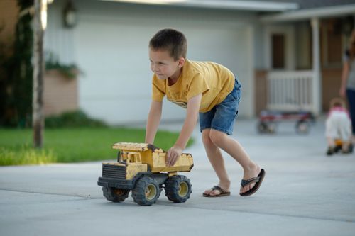 A boy in a yellow shirt, shorts, and flip flops pushes around a yellow toy dump truck in a driveway.
