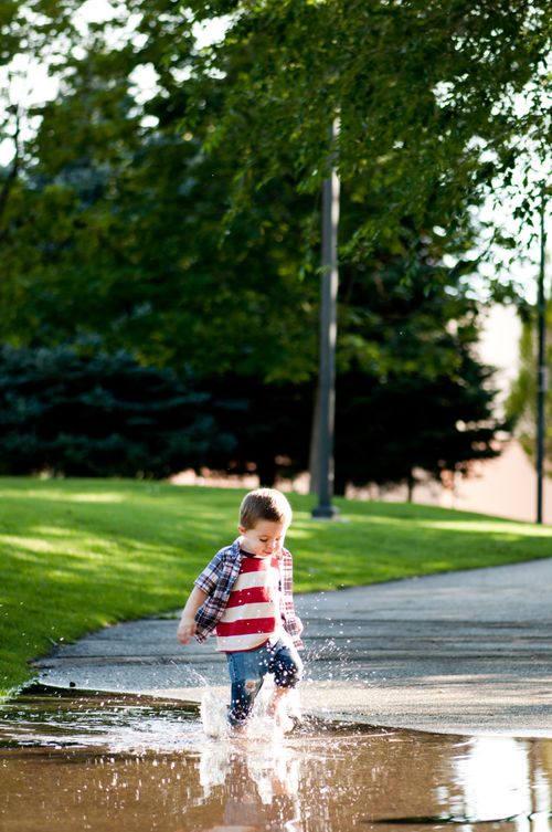 A little boy in an American-flag shirt and blue jeans splashes his feet in a puddle of water on the road.