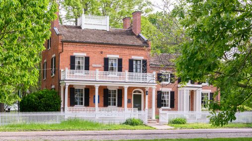 A two-and-a-half story brick home with a double white porch and sizable addition to the right with a white picket fence.