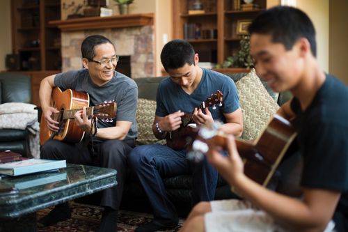 A father sits on a couch and plays a guitar beside his two sons, who are playing a ukulele and guitar.