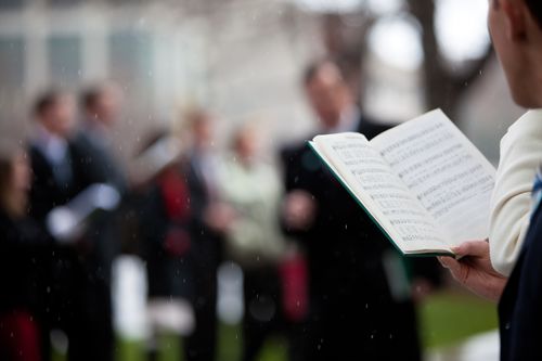 A close-up image of a man standing and holding a hymnbook while caroling with others in the snow.