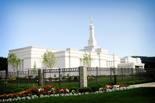 The entire Spokane Washington Temple during the daytime, with grounds surrounded by a fence, lined with flowers and trees.