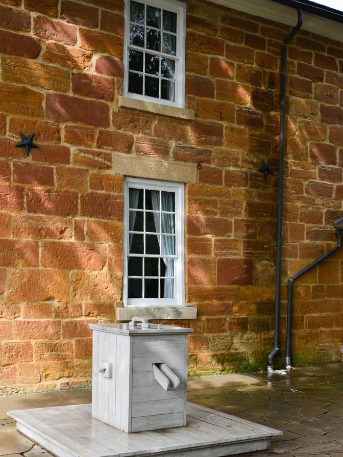 A water pump made of gray wood on a small wooden platform beneath a second-story window.