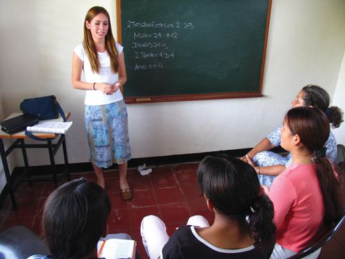 A young woman with long brown hair and in a skirt stands at the front of a classroom next to a blackboard and teaches to a room of five young women.