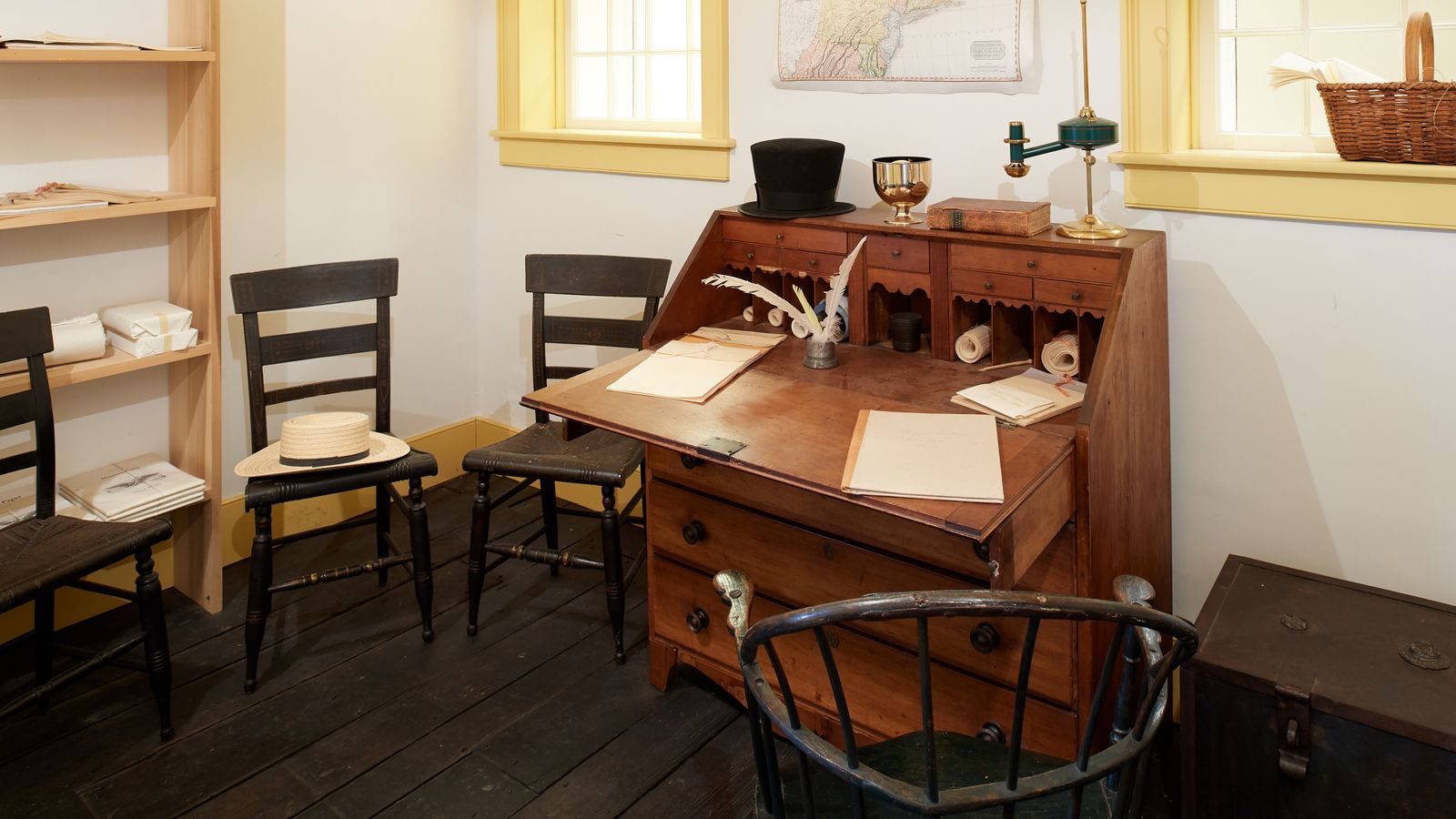 We see interiors of a building. It could be the Grandin print shop but it is unclear. These images include a wooden writing desk up against a wall with wooden chairs near by.