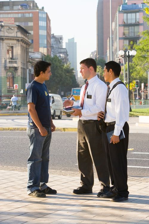 Two male missionaries in white shirts and ties standing on a busy street corner, talking to a young man in jeans and a T-shirt.