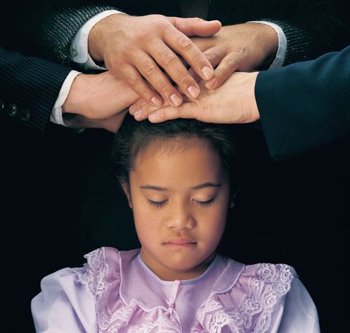 A young black-haired girl in a purple dress closes her eyes while three men place their hands on her head, confirming her a member of the Church.