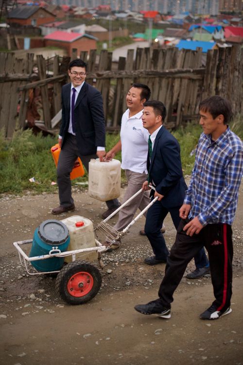 Two ministering brothers in suits help two men carry jugs and push a cart down a dirt road next to a wooden fence.