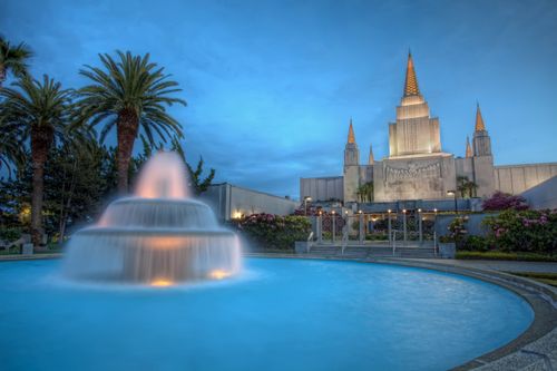 A water fountain on the grounds of the Oakland California Temple, with the temple seen lit up in the background during the evening.