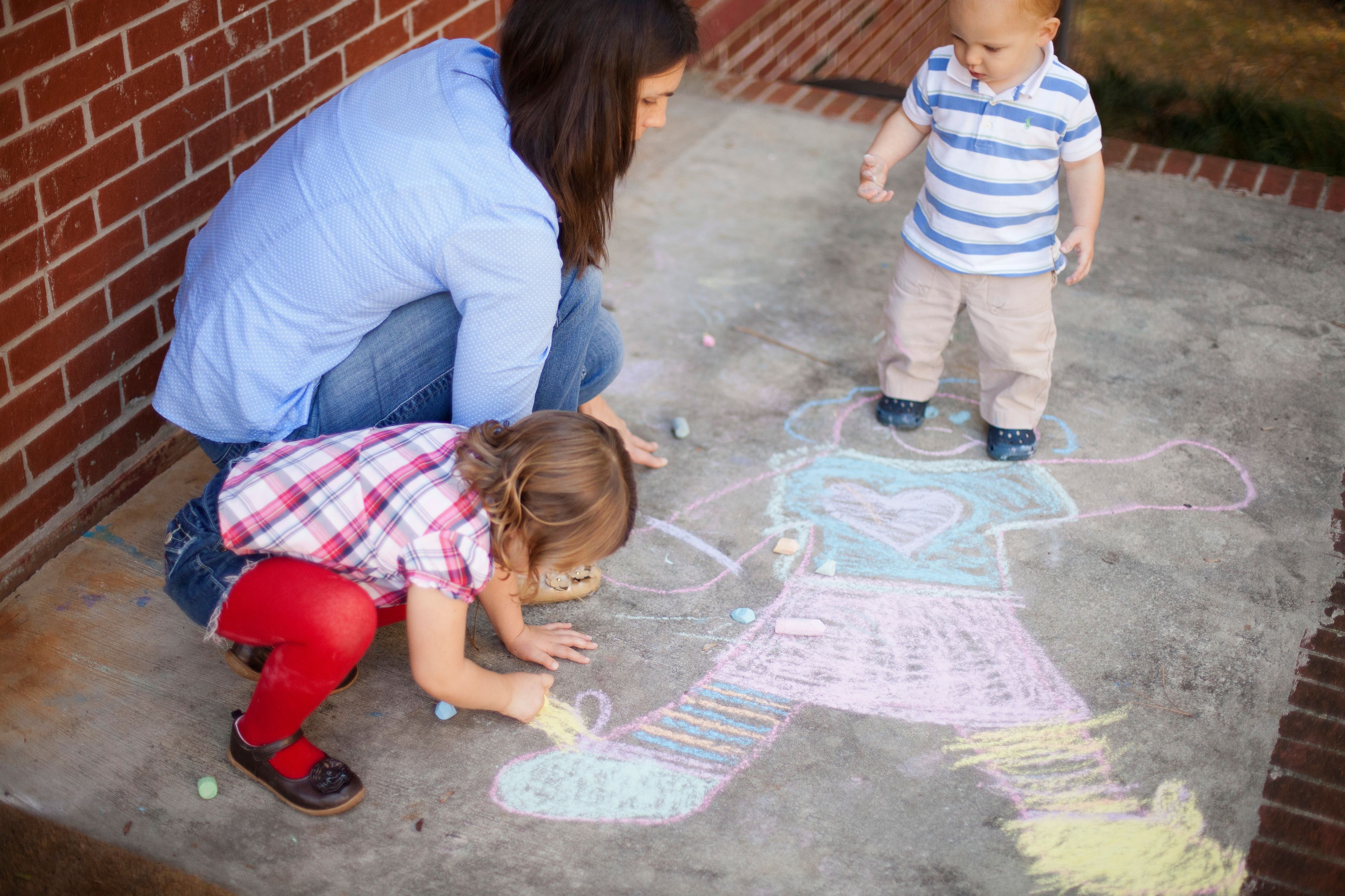 A mother kneels on the ground beside her son and daughter, who are drawing with chalk.