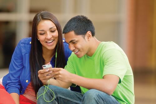 A young woman and young man share a set of headphones to listen to music from an iPod together while sitting down outside.