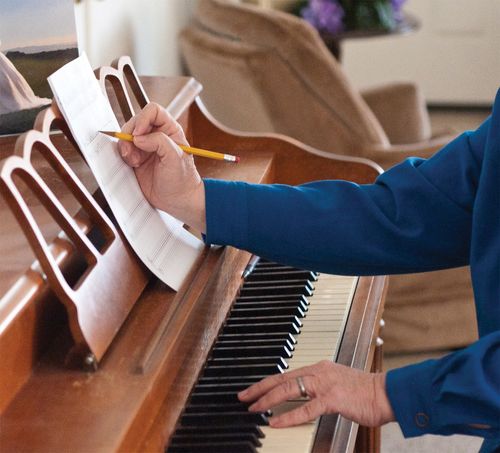 A woman sits at a piano and touches the keys with one hand and makes notes on her sheet of music with the other.