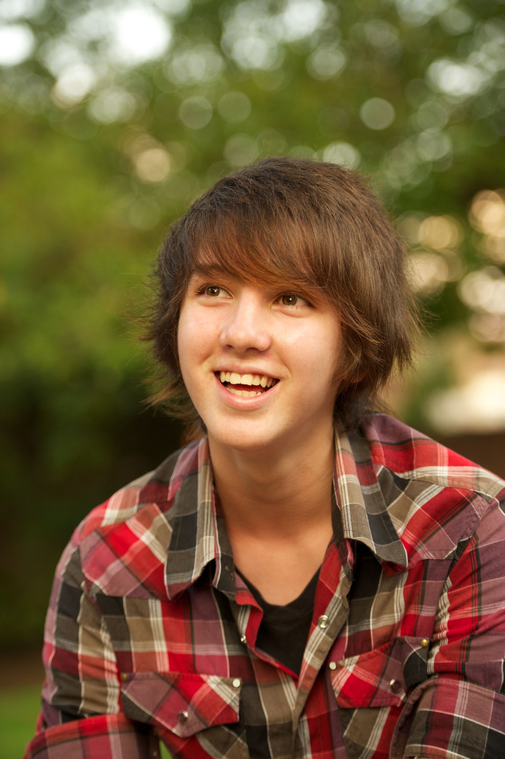 A portrait of a young man with somewhat long hair and a plaid shirt, looking up and smiling.