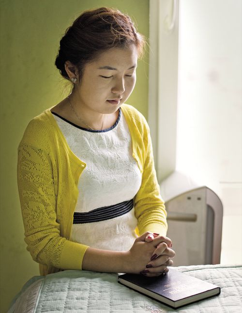 A young woman kneeling by her bed in prayer.  There is a copy of the Book of Mormon on the bed in front of her.