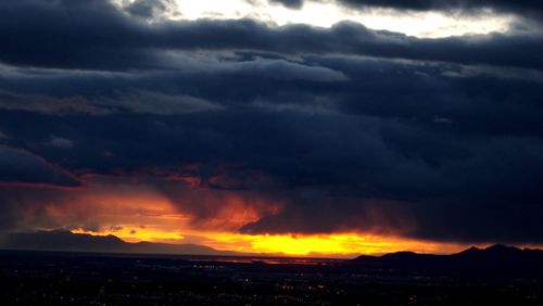 Dark storm clouds gather over a valley while the sun sets in the distance, reflecting deep orange and yellow sunlight.