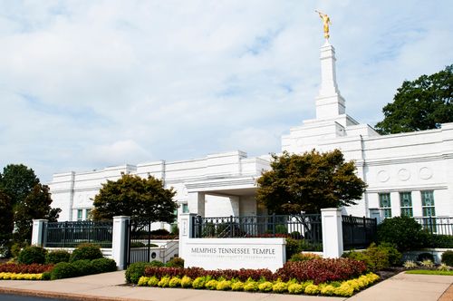 The Memphis Tennessee Temple sign surrounded by flowers, with the temple in the background and clouds overhead.