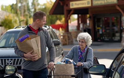 A man helping an elderly woman with her groceries.  They are in a parking lot.
