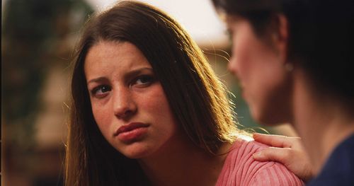 A young woman sitting down. A woman (possibly her mother) has her hand on the girl’s shoulder. The girl has a sad expression on her face.