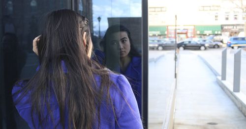 Eine Frau betrachtet ihr Spiegelbild im Fenster eines Gebäudes; im Hintergrund eine Straße