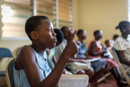 A young woman raises her hand during Sunday School with her scriptures open on her lap and other students sitting around her in the classroom.