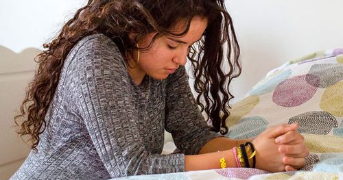 A young woman kneels by her bedside to pray.