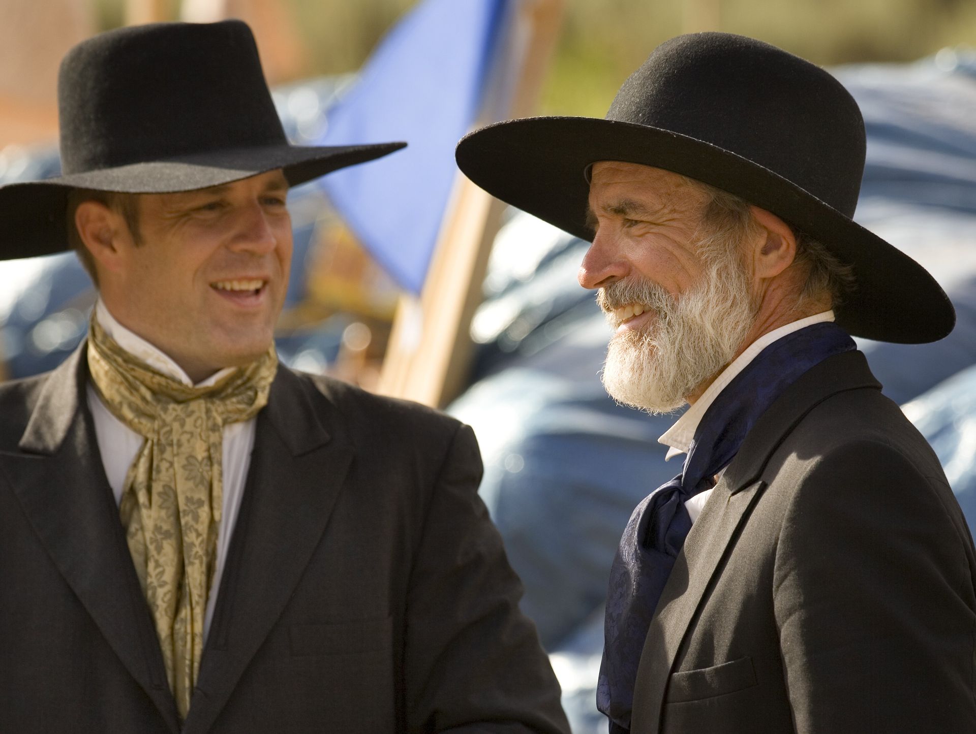 Two men dressed in suits and wide-brimmed hats stand and talk together.