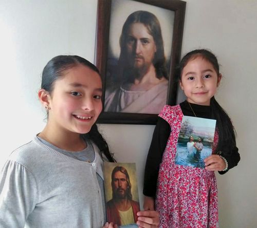 Two young sisters both holding different pictures of the Savior while standing in front of a picture of the Savior