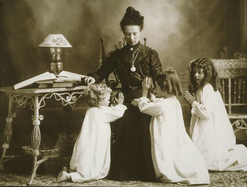 George Edward Anderson photograph of Mrs. Albert Manwaring and her children. Mrs. Manwaring is wearing a black dress and is seated in a chair. Three young girls in white gowns are kneeling beside their mother. The children have their hands clasped in preparation for prayer. Mrs. Manwaring has her head bowed. The original photograph was taken in Springville, Utah in 1903. Mrs. Manwaring sent the photograph to her husband Albert while he was serving an LDS mission in England.
