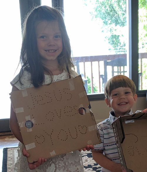 A young girl and a young boy hold cardboard with holes punched in it. The girl's has Jesus Loves You punched into it.