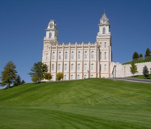 A side view of the Manti Utah Temple on top of a green grassy hill with trees surrounding.