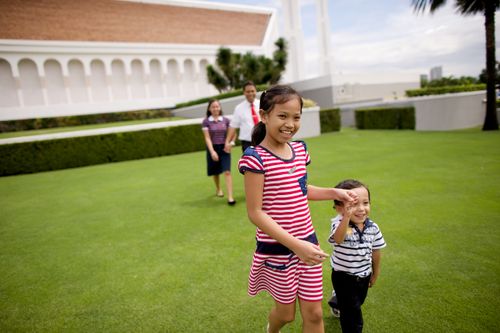 A girl walking and holding hands with her younger brother as their parents walk behind them, holding hands, outside the Manila Philippines Temple.