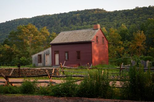 Dark red clapboard house with a lighter annex and a rail fence in the foreground..