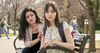 two young adult women sit on a park bench; one looks sad, and the other is reaching to comfort her