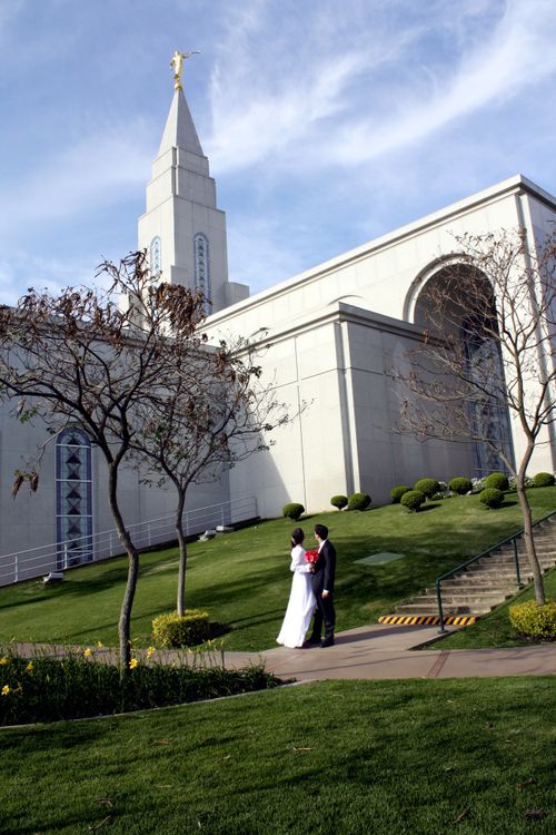 A bride holding a bouquet of red flowers and standing beside the groom as they look up at the Campinas Brazil Temple.